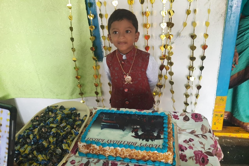 A little boy in a vest standing behind a cake reading "happy birthday Krish"