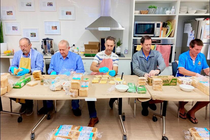 Five men sit along a table making sandwiches