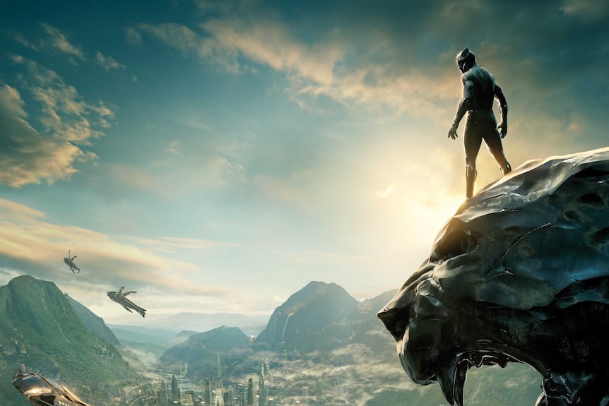 Still image from 2018 film Black Panther of the main character standing atop a panther rock face looking over a city.