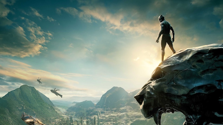 Still image from 2018 film Black Panther of the main character standing atop a panther rock face looking over a city.