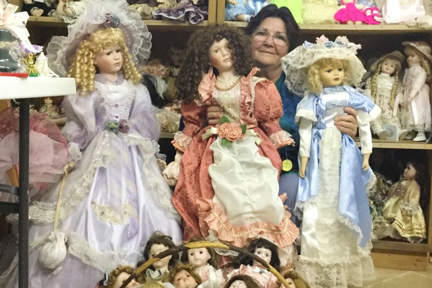 Woman in centre of three big dolls with basket of dolls in foreground and bookcase of dolls in background