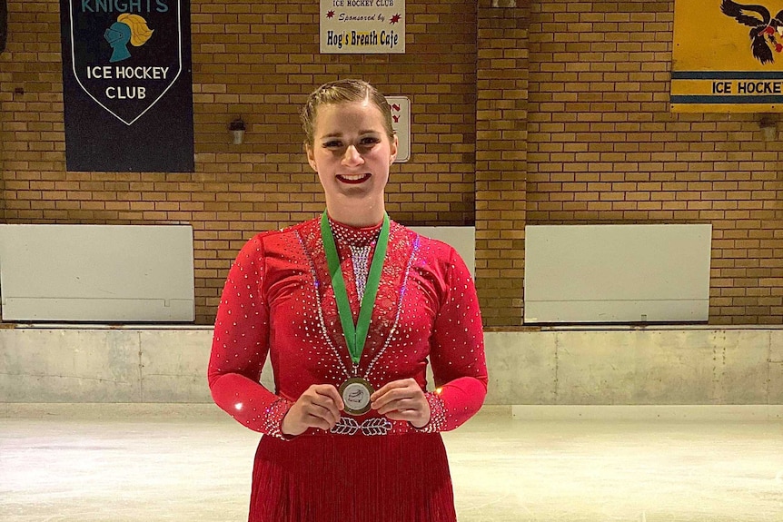 An ice skater in a red dress stands on the ice holding a medal.