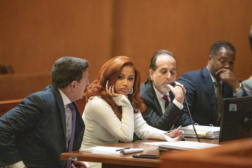 A black woman with long red hair and long white fingernails sits at a table in a courtroom listening to a male lawyer.