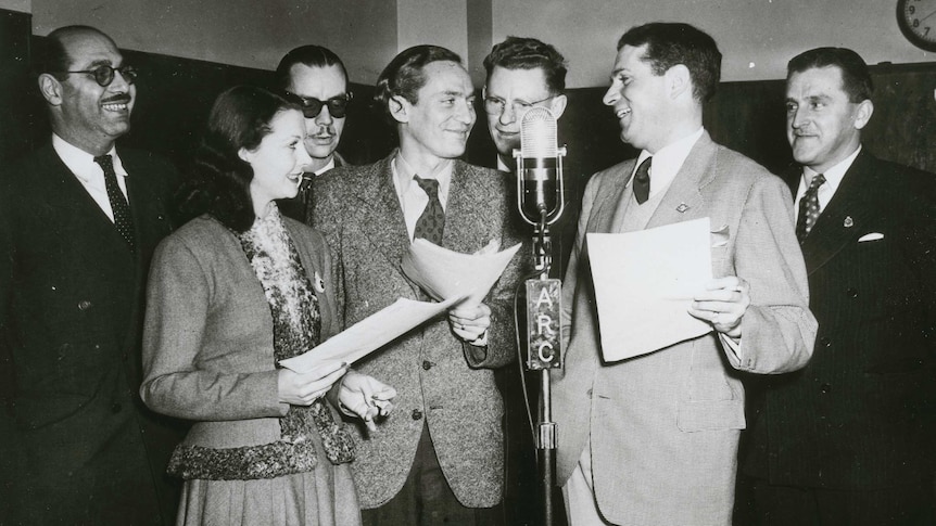 Vivien Leigh, Peter Finch & Laurence Olivier stand around a microphone smiling at each other, flanked by 4 smiling men in suits.
