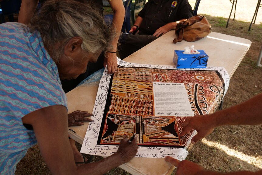 A woman leaning down and signing a large document featuring traditional Indigenous artwork, on top of a plastic trestle table.  
