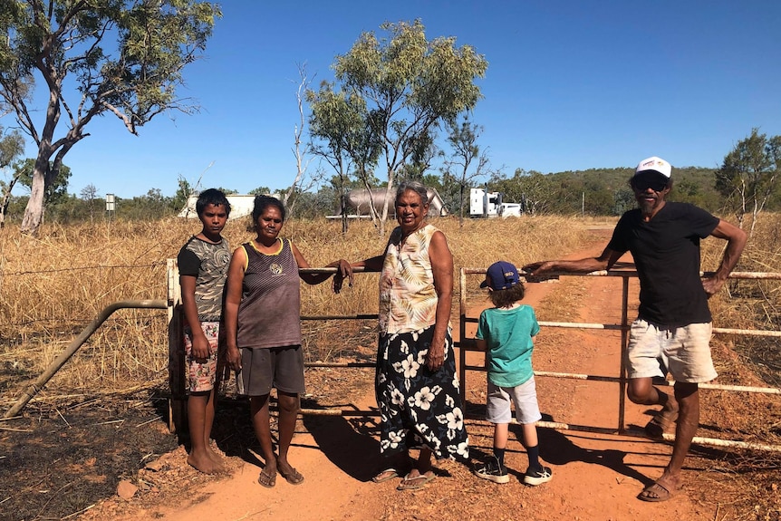 A family of five stand on a dusty driveway in front of a metal gate.