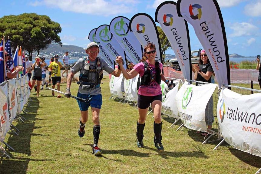 An elderly man and a middle aged woman run across a finish line hand in hand.