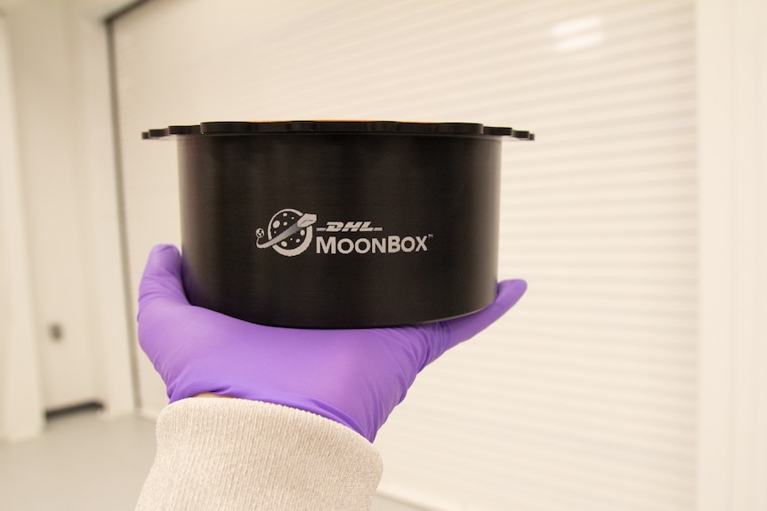 Black circular box with MoonBox written on it, purple glove holding it. 
