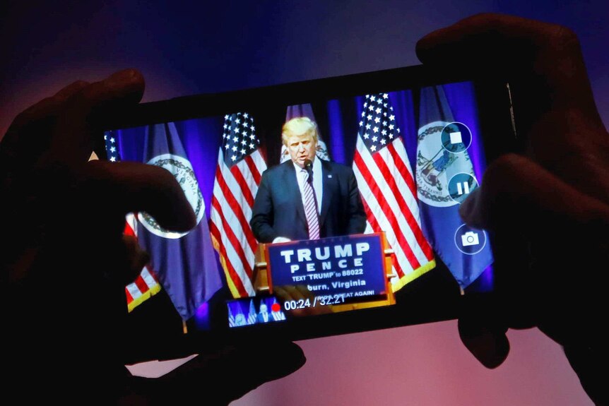 Hands holding a mobile phone with an image of Donald Trump on it.