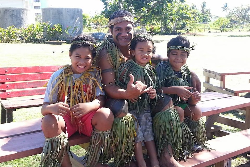 A young boy sits on a bench with a man and two small children. They are all dressed in garments made of grass.