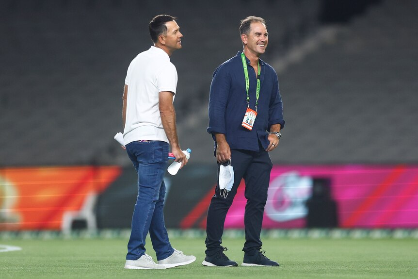 Ricky Ponting and Justin Langer, wearing casual clothes, smile and chat in the middle of the field