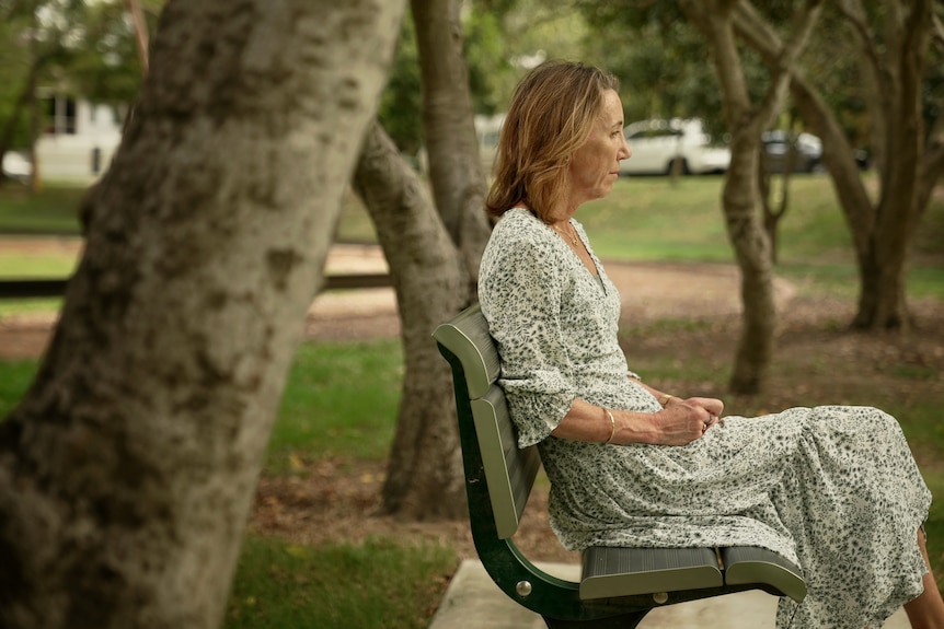 A woman in a dress sits on a park bench staring ahead. 