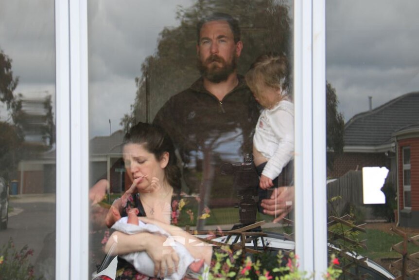 A man holds a toddler and a woman sits holding a newborn. They are in their home, the photo is take through the window.