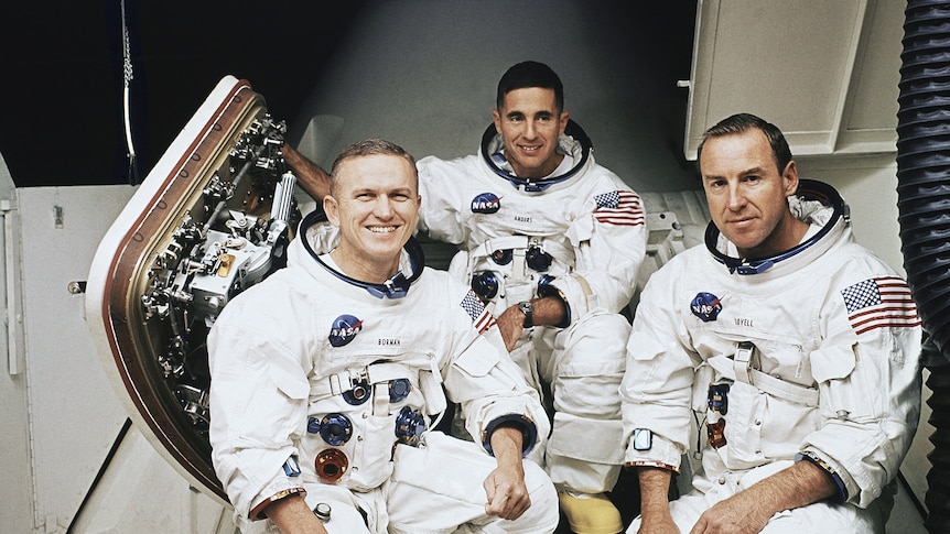 Three men in space suits, crouch down while smiling.