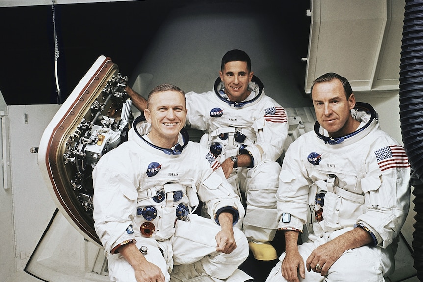 Three men in space suits, crouch down while smiling.