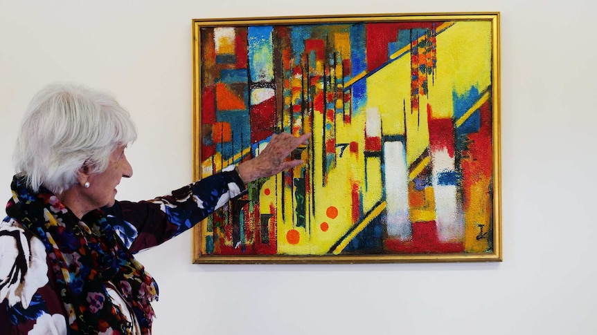 A senior woman pointing to her artwork which is an abstract, colourful piece in a golden frame hanging on a white wall.