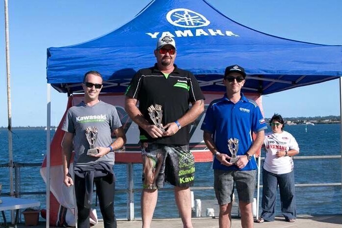 Cameron Martin (centre), in sunglasses and cap, stands on the first place spot on a podium, holding a trophy