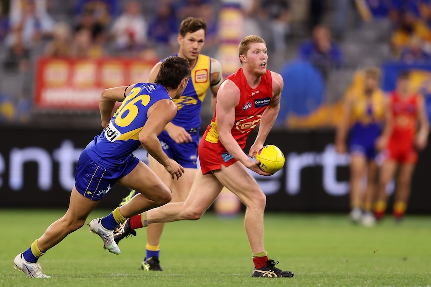 A Gold Coast AFL player looks up while moving his fist to deliver a handball while opponents try to close in on him.