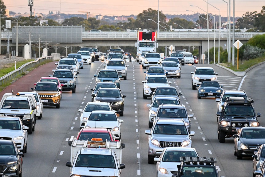 A mix of cars and trucks fill four lanes of peak hour traffic on freeway.