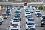 A mix of cars and trucks fill four lanes of peak hour traffic on freeway.
