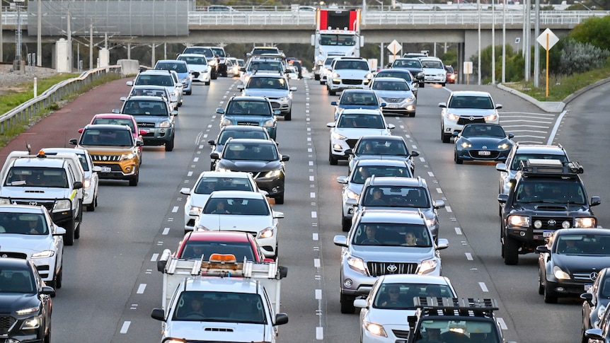 A mix of cars and trucks fill four lanes of peak hour traffic on freeway.