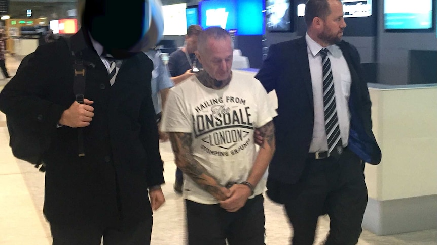 A man in t-shirt and thongs is escorted by policemen.