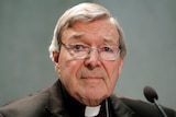 A headshot of Cardinal George Pell, with a stand microphone in the bottom right corner. He is wearing silver-rimmed glasses.