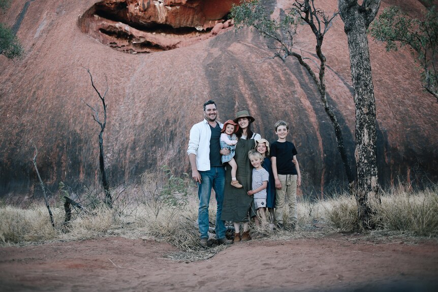 Against a large red rock, a man and woman and four young children, wearing neat casual clothes, stand close together smiling.