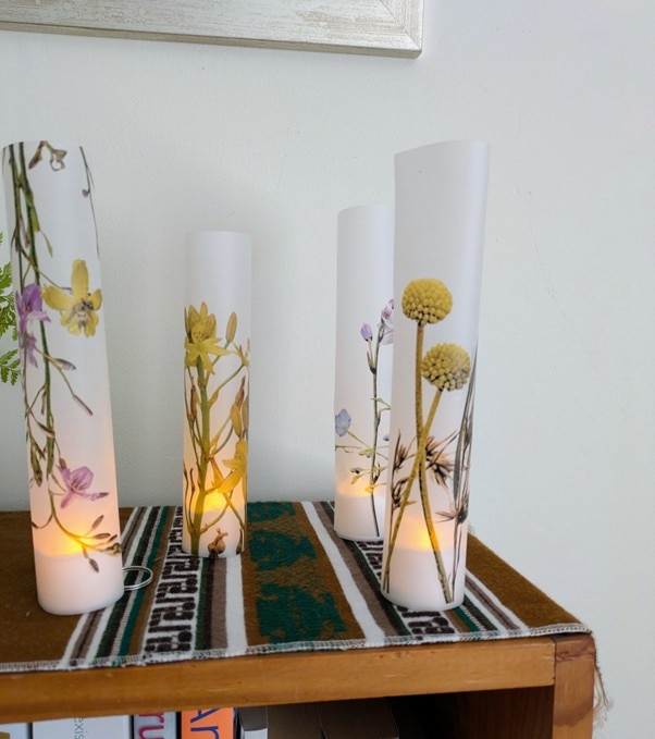 Quatro velas altas brancas com imagens de flores nativas.