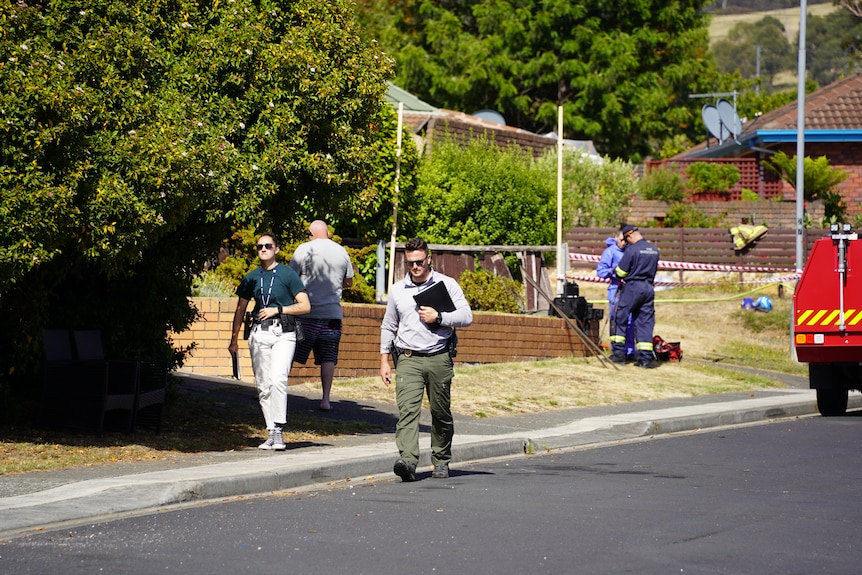 Two police detectives wearing casual clothes walk along a suburban footpath. Forensic officers are visible in the background