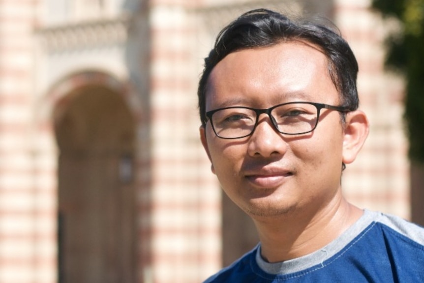 An Asian man wearing dark-rimmed glasses and a royal blue T-shirt stands in front of a sandstone building.