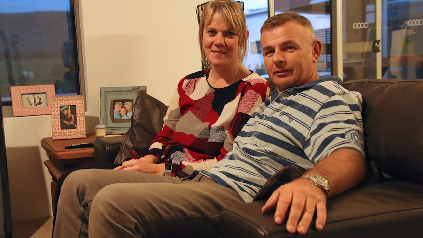 Sandra and Mark sit on a couch with pictures of their kids in the backgorund.