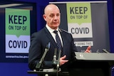 Tasmanian Premier Peter Gutwein in a suit gestures at a press conference podium in front of a "Keep on top of COVID" sign.