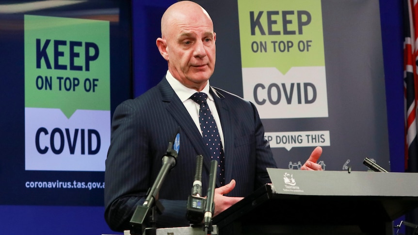 Le Premier ministre tasmanien Peter Gutwein en costume fait des gestes lors d'une conférence de presse podium devant un "Restez à l'affût de COVID" signe.