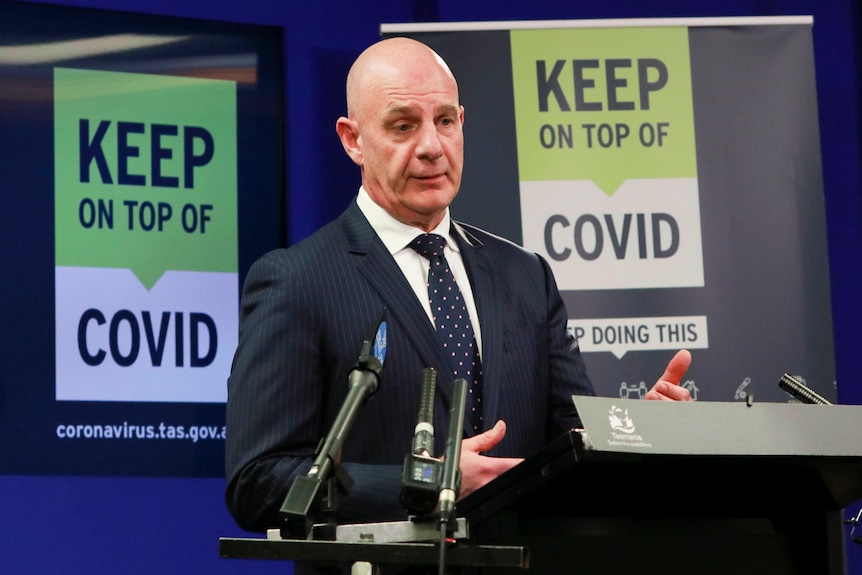 Tasmanian Premier Peter Gutwein in a suit gestures at a press conference podium in front of a "Keep on top of COVID" sign.