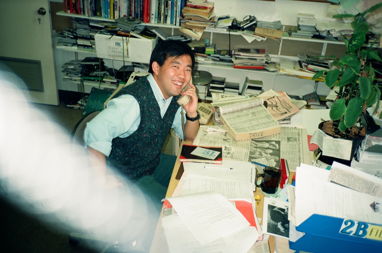a man smiling while using a landline at a desk covered in newspapers