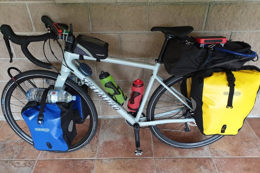 A bicycle leans against a brick wall with a large yellow pack attached to the back wheel and a blue pack attached to the front.