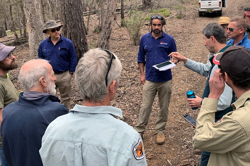 A group of men dressed in work uniforms stand in a forest conferring with one another.