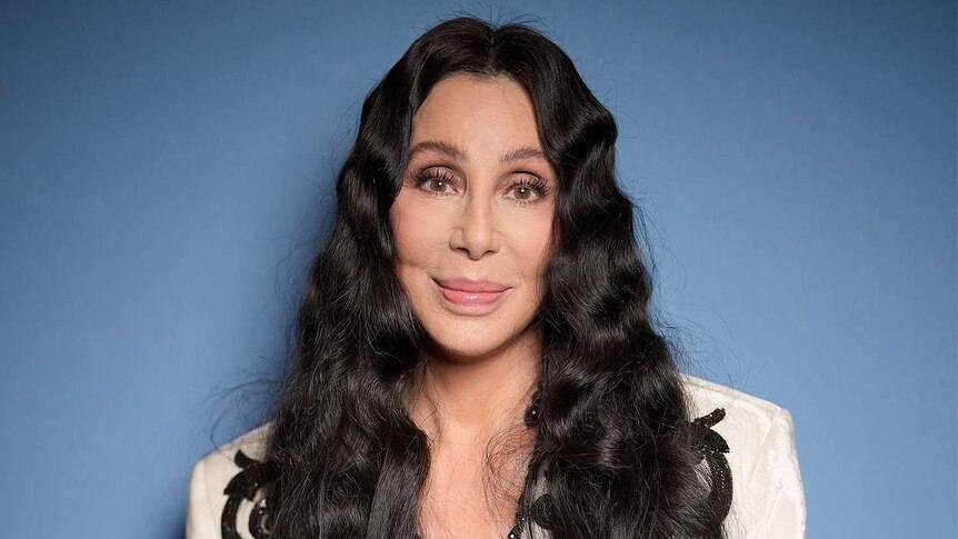 closeup of Cher with black wavy hair looking into the camera wearing a while jacket against a blue background