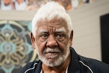 A man with white hair and facial hair looks seriously at the camera. Behind him is an Indigenous print.