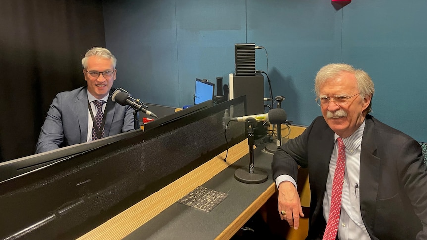 2 men in a radio studio look toward the camera with microphones on a desk 