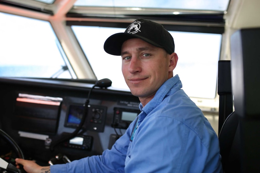 A man in a blue shirt and black baseball cap in the cabin of a ship.