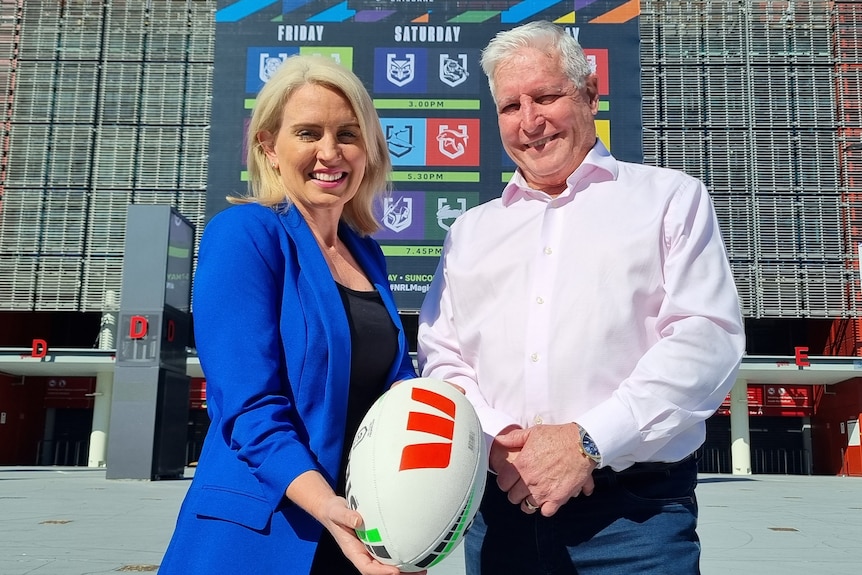 A woman in blue and a man in pink hold a footy in front of Lang Park stadium