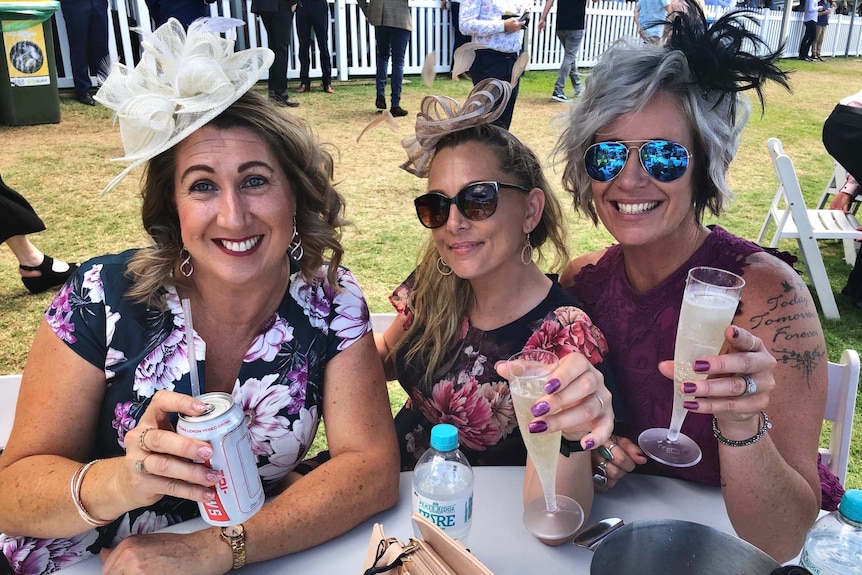 Three women with sparkling wine and a can of vodka posing at a table wearing fascinators