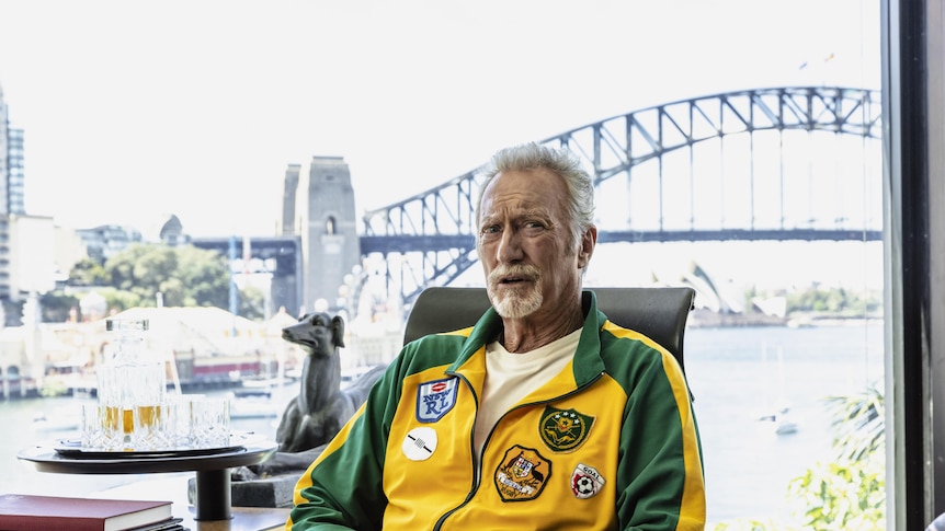 Bryan Brown dressed in a green and gold tracksuit sitting in an office, the Sydney Harbour Bridge behind him