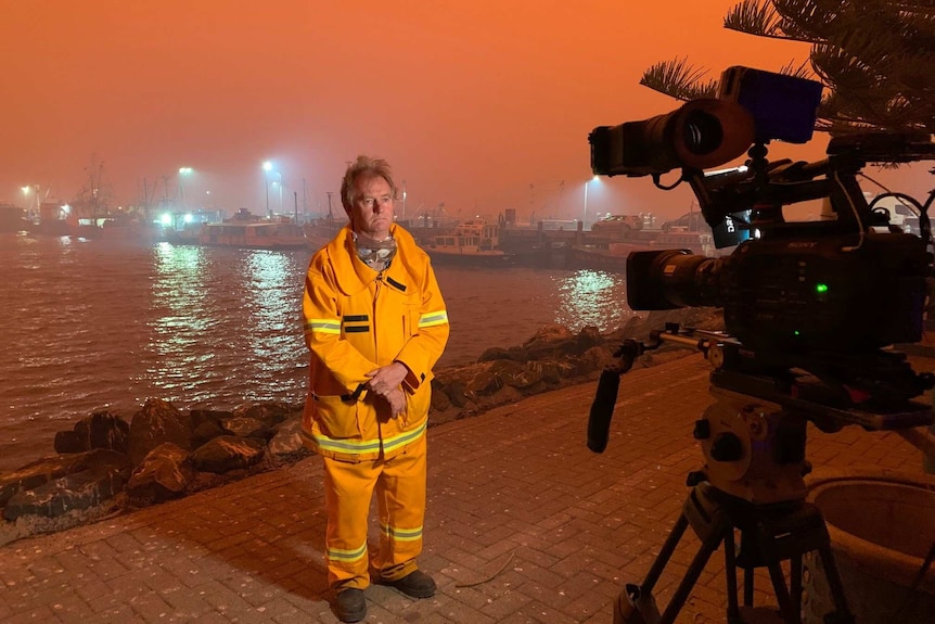 Williams standing in front of TV camera near wharf with smoky red sky over water.