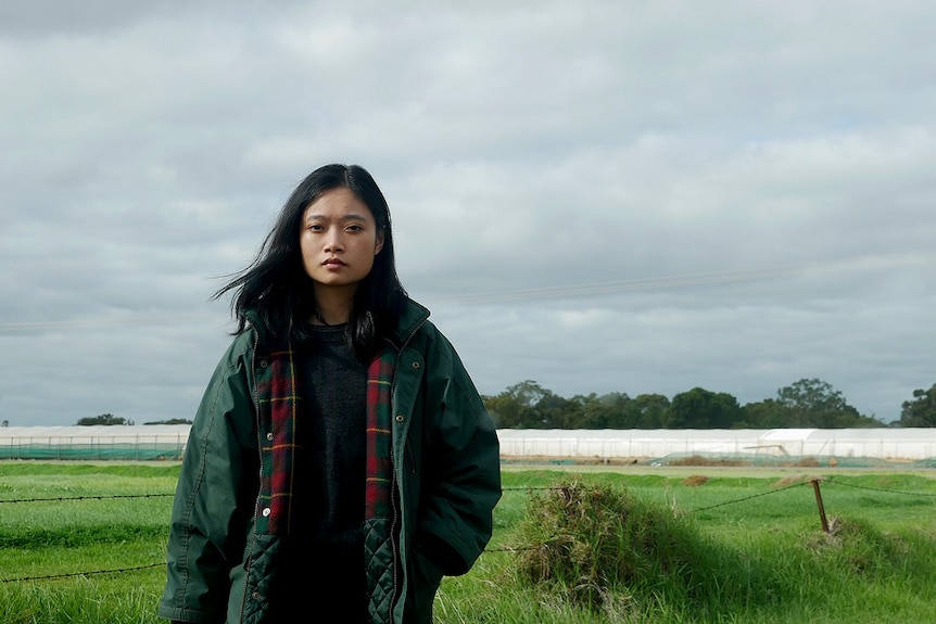 A woman stands looking at the camera with a greenhouse in the background.