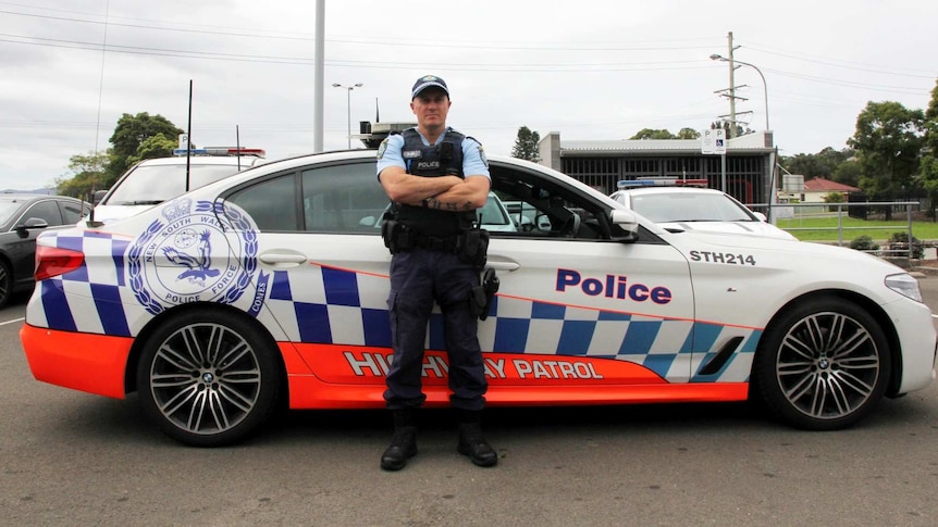 A highway patrol policeman stands in front of his cruiser with his arms folded.