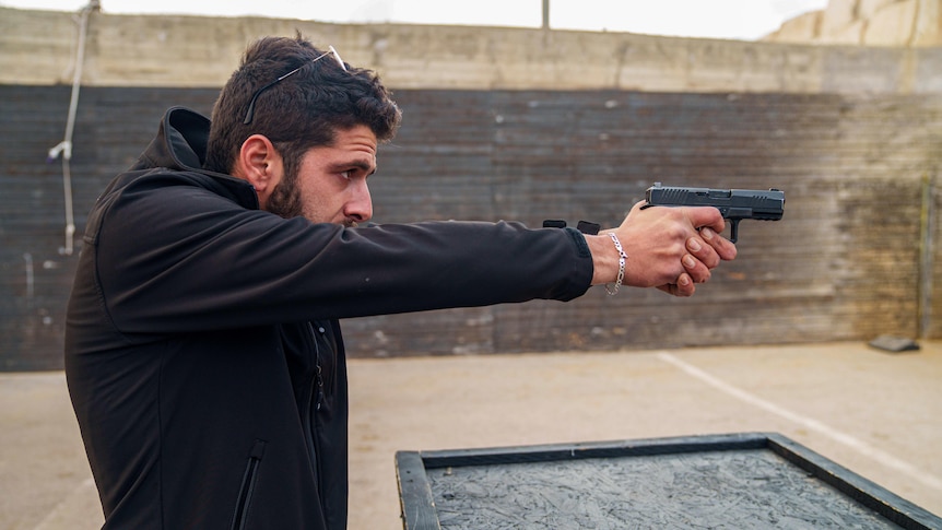 A man wearing a black jacket holds a pistol with two hands, outstretched in front of him