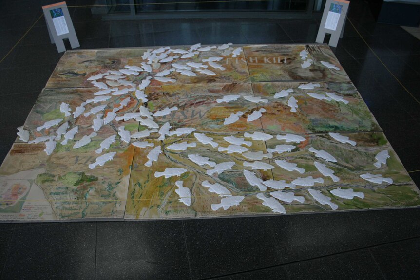 A large mat lies on the ground with a hand-drawn map of Menindee and about 100 3D printed fish placed on top